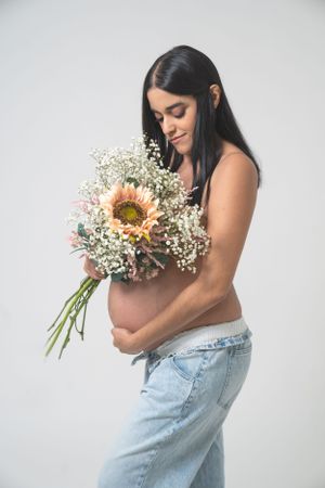 Pregnant woman in casual attire holds delicate bouquet of sunflowers and baby's breath.