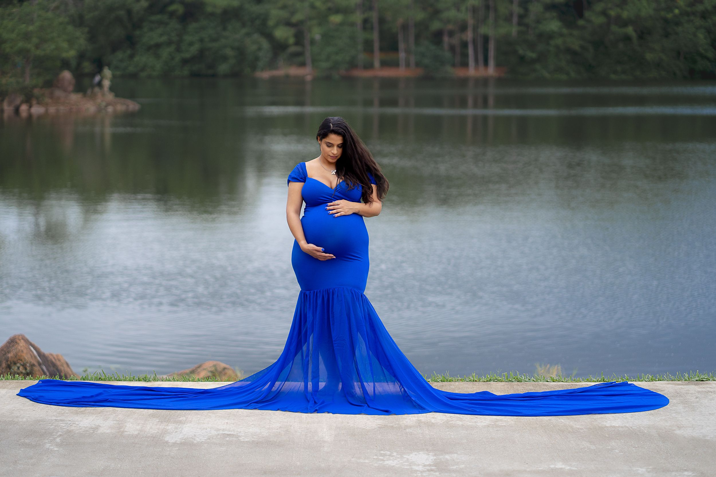 Maternity portrait in royal blue flowing gown by tranquil lake.