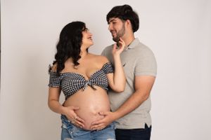 Expecting couple poses together showing pregnant belly in casual attire against neutral background.