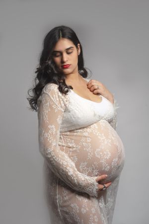 Pregnant woman poses in white lace maternity dress against grey background.