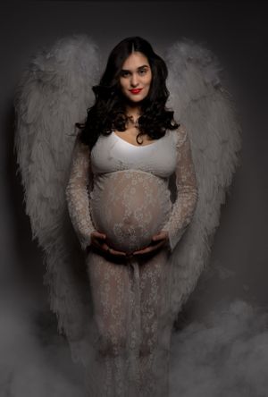 Ethereal maternity portrait with angel wings and lace dress against dark background.