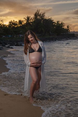 Sunset maternity photo of woman in bikini and coverup on beach.