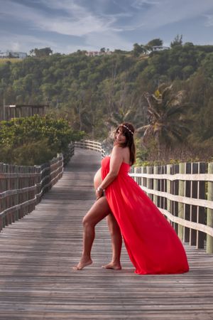 A flowing red dress catches the wind on a wooden boardwalk surrounded by lush tropical foliage.