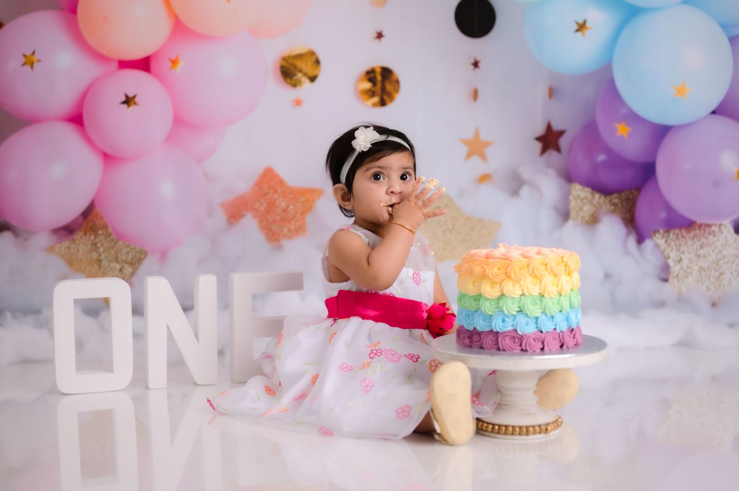 Colorful balloon arch and cake setup for a first birthday celebration with pastel pink and blue decorations.