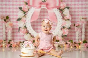 Pink gingham backdrop with floral decorations and birthday cake setup in soft lighting.