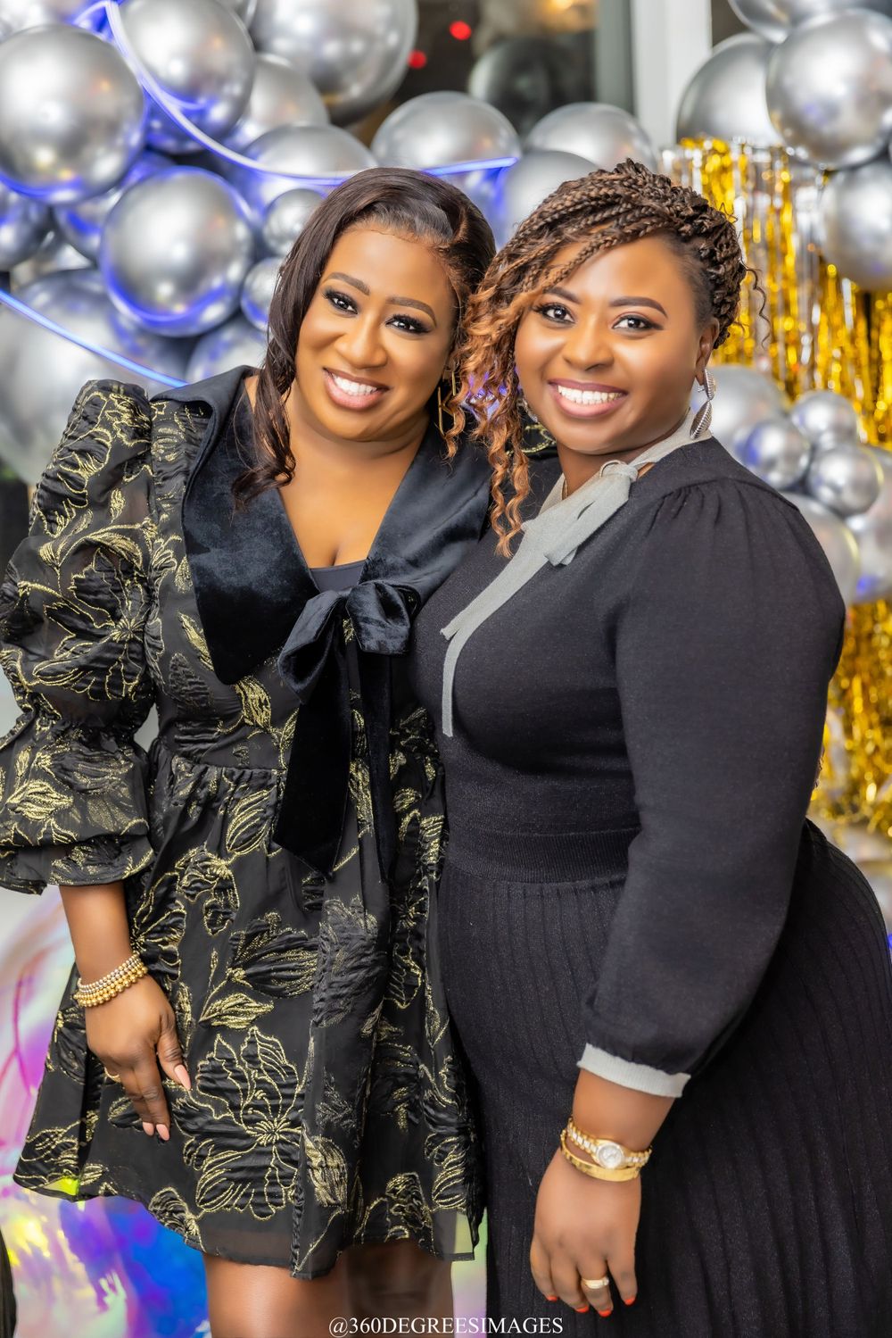 Two smiling women in black outfits pose together at a celebration with silver and gold balloon decor.