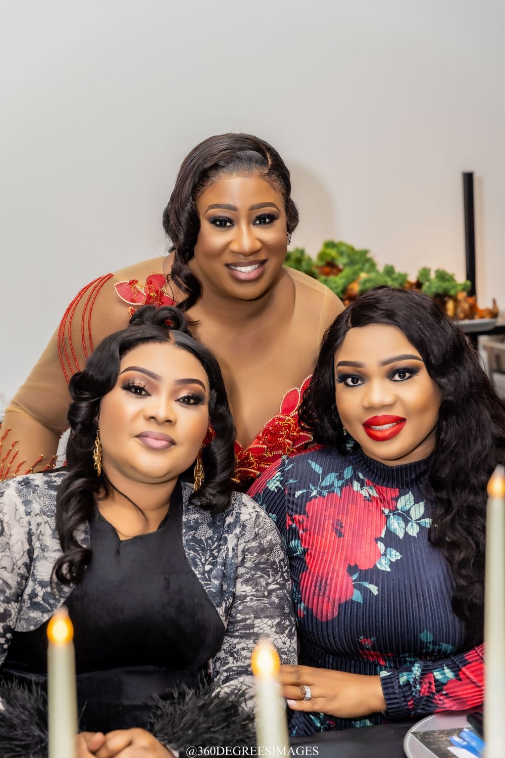 Three elegantly dressed women pose together at a candlelit event, smiling warmly for the camera.