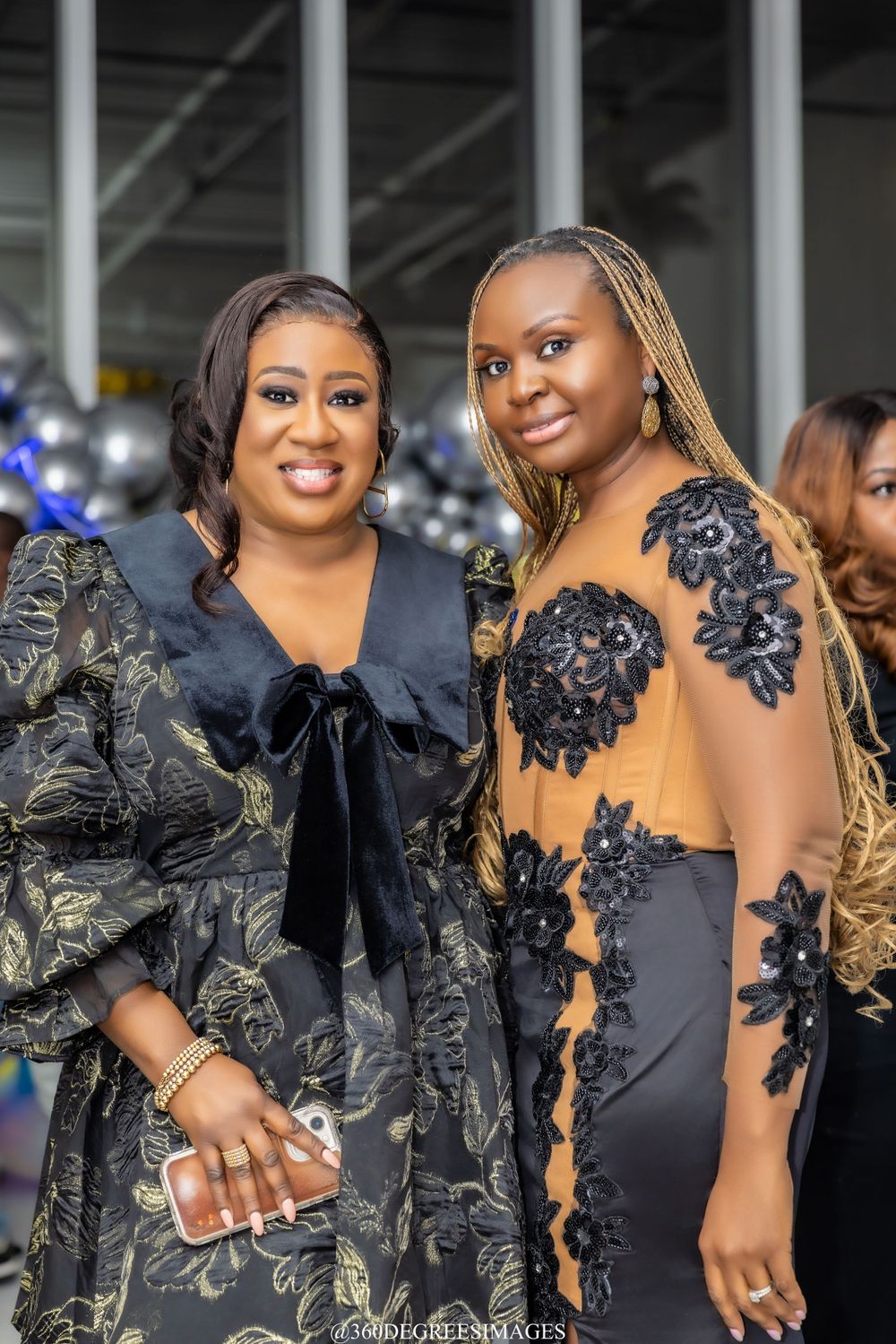Two stylish women in black outfits with floral embellishments pose together at an elegant celebration event.