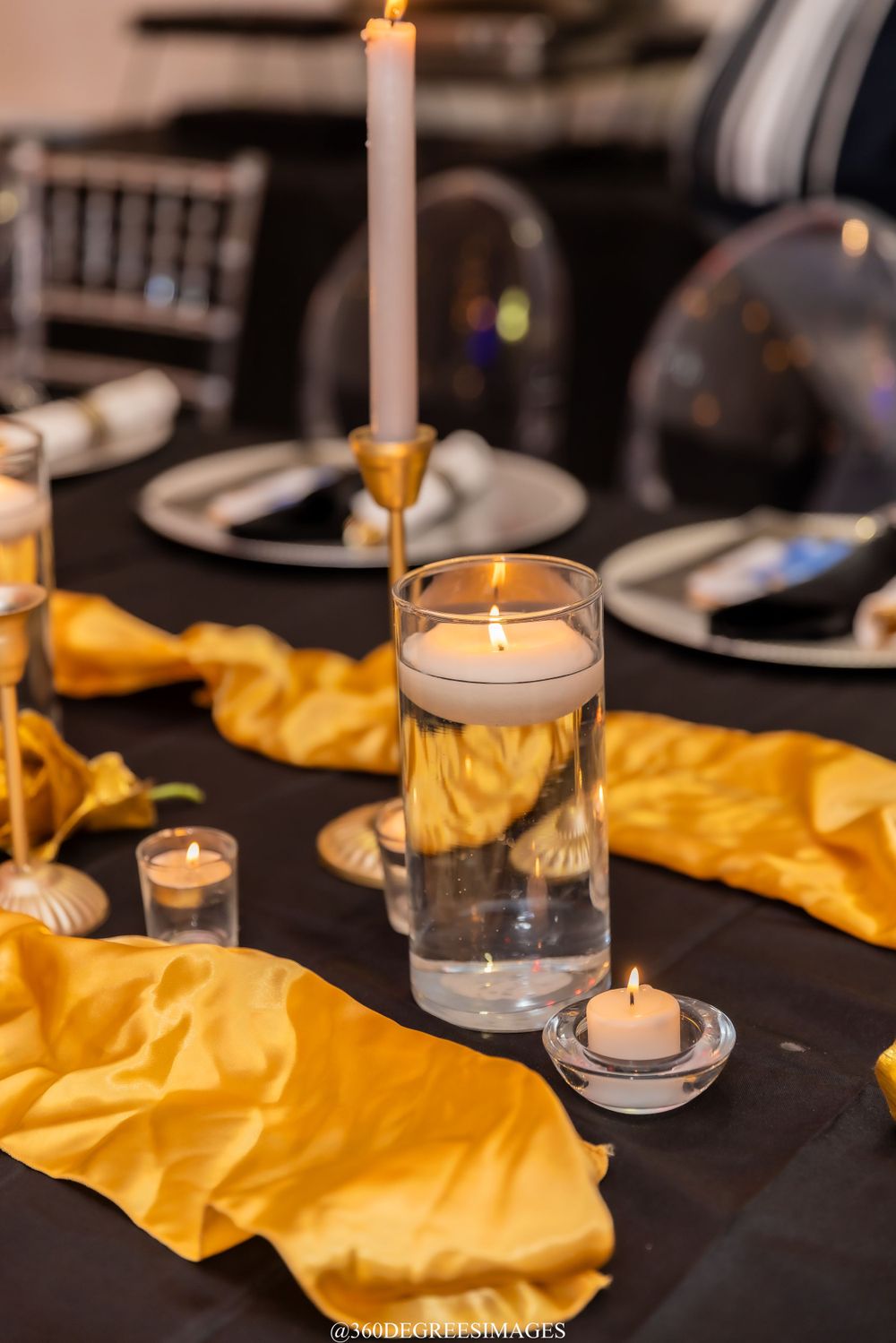 Black tablecloth with gold satin runner, floating candle centerpiece, taper candles, and silver charger plates.