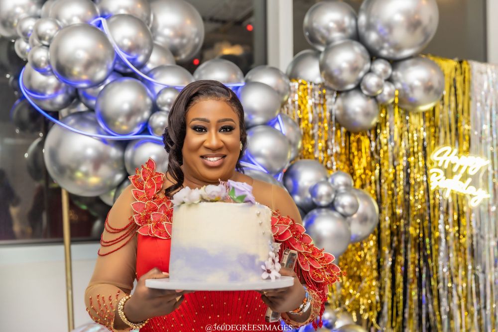 Smiling woman in red lace outfit holds white birthday cake at festive party with silver balloon decor.
