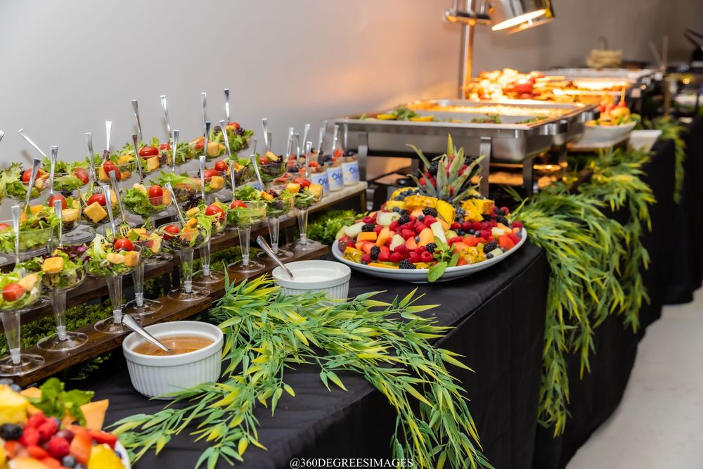 Catered buffet table featuring fresh fruit platters, salad cups, and lush green garland decorations.