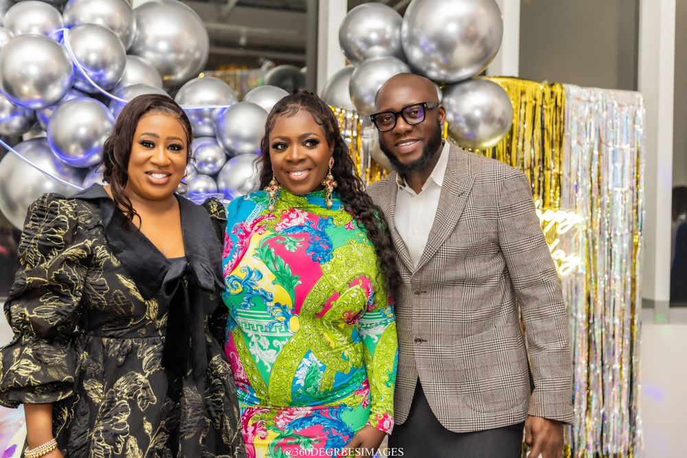 Three guests smile together at a festive event decorated with silver and gold balloons in the background.