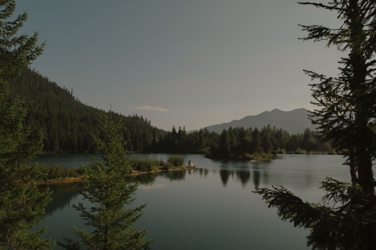 Serene mountain lake surrounded by evergreen trees reflects the cloudy sky at dusk.