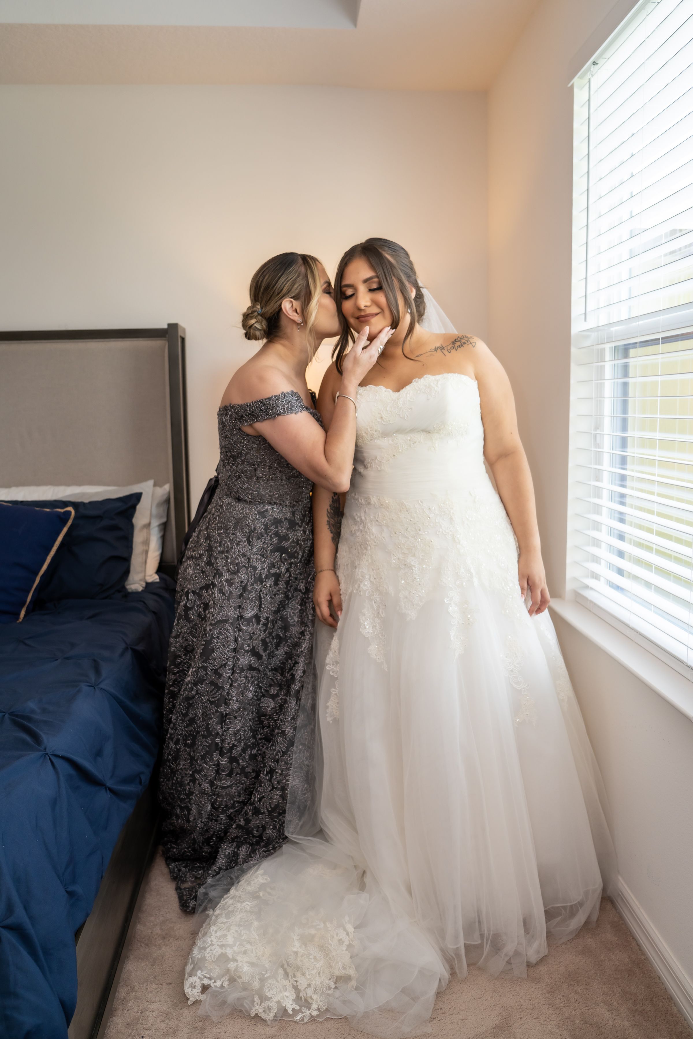 Two people share an intimate moment while preparing for a wedding, one wearing a white gown.