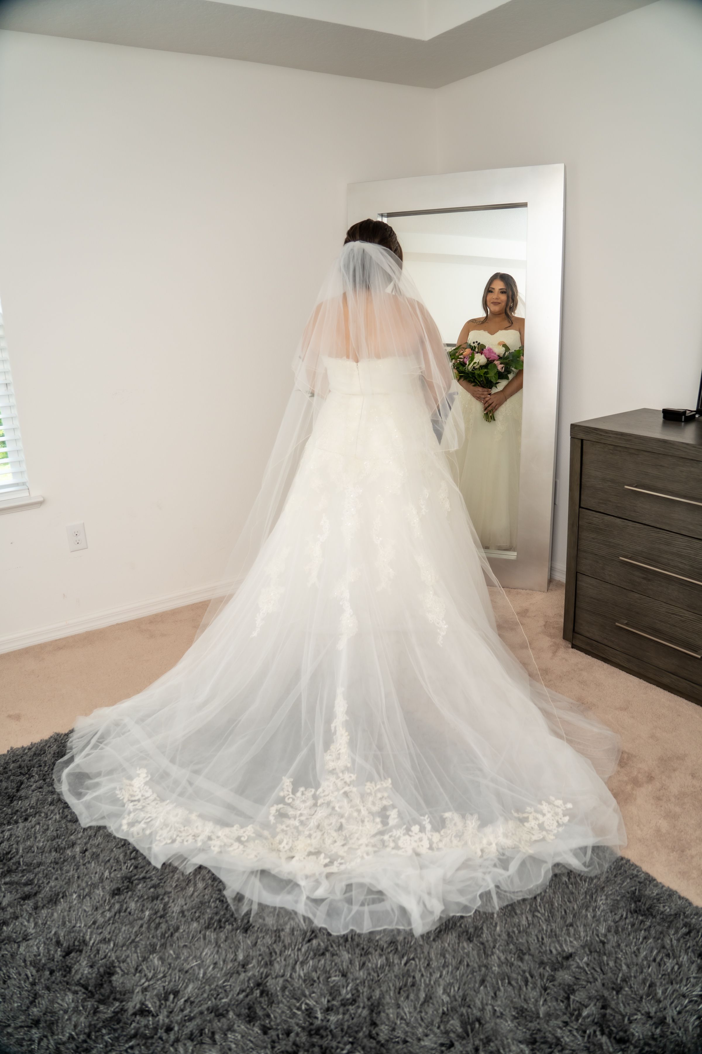 A bride in a flowing white wedding gown with long train looks at her reflection in a full-length mirror.