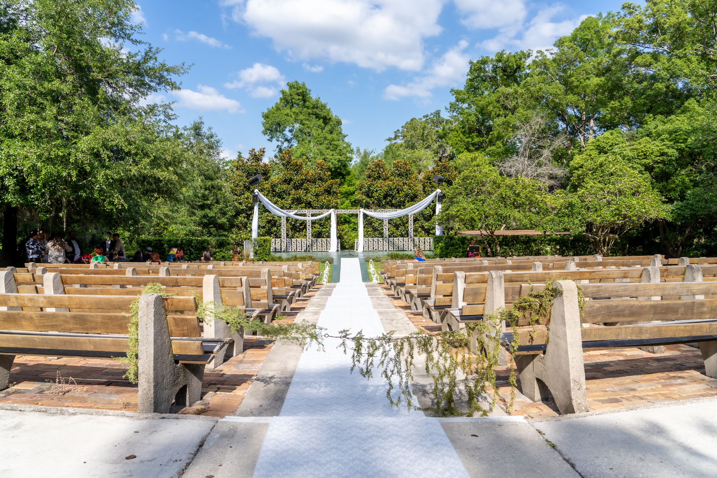White wedding ceremony setup with floral arrangements and wooden benches under blue sky and trees.