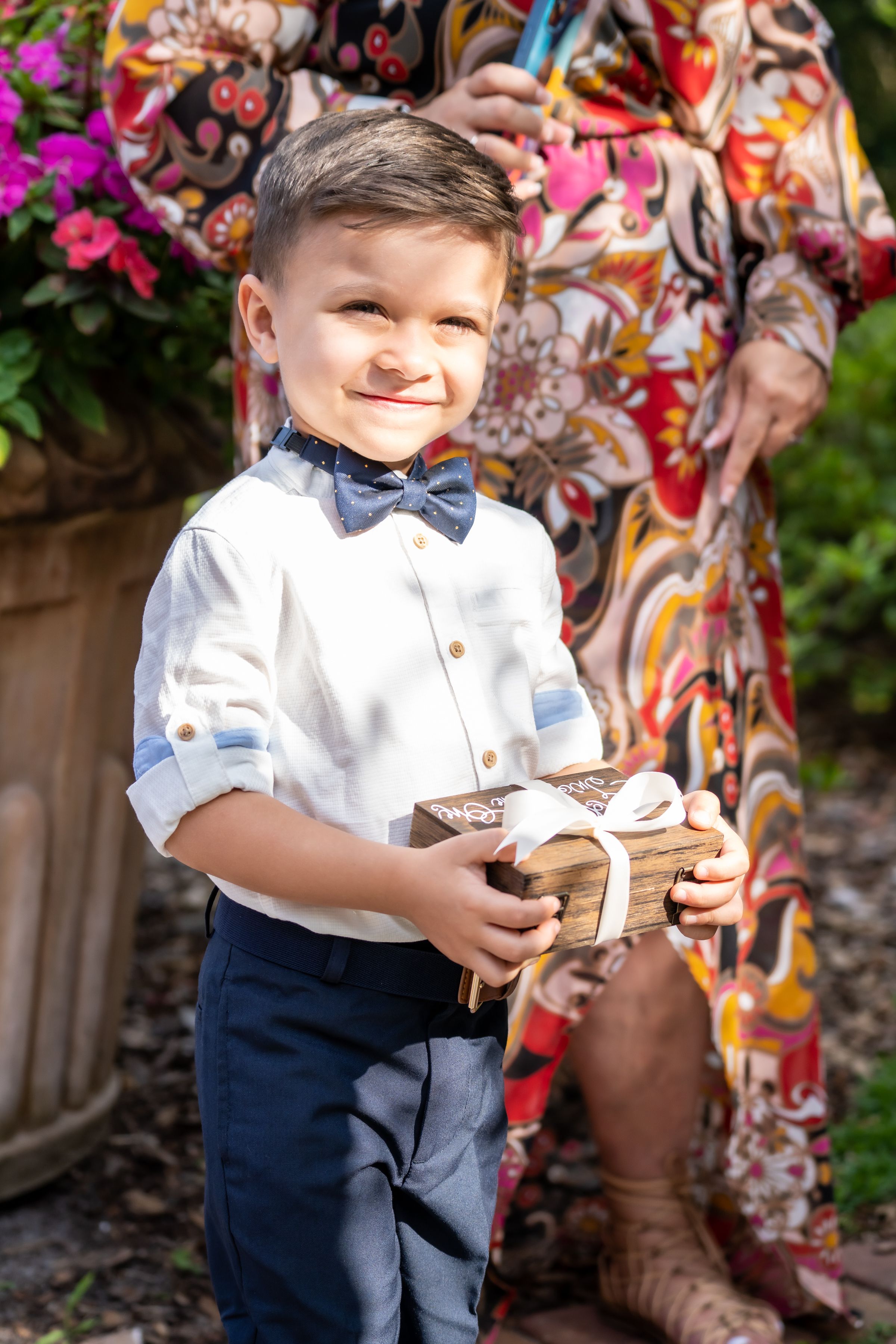 A young boy in formal attire with bow tie holds a gift box while standing near colorful flowers.