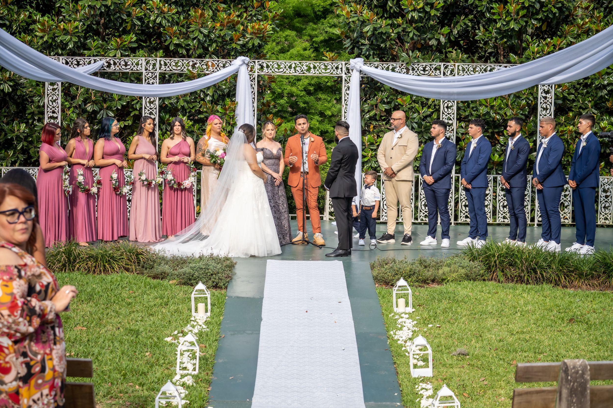 An outdoor wedding ceremony setup with bridesmaids and groomsmen standing under a white draped arbor.