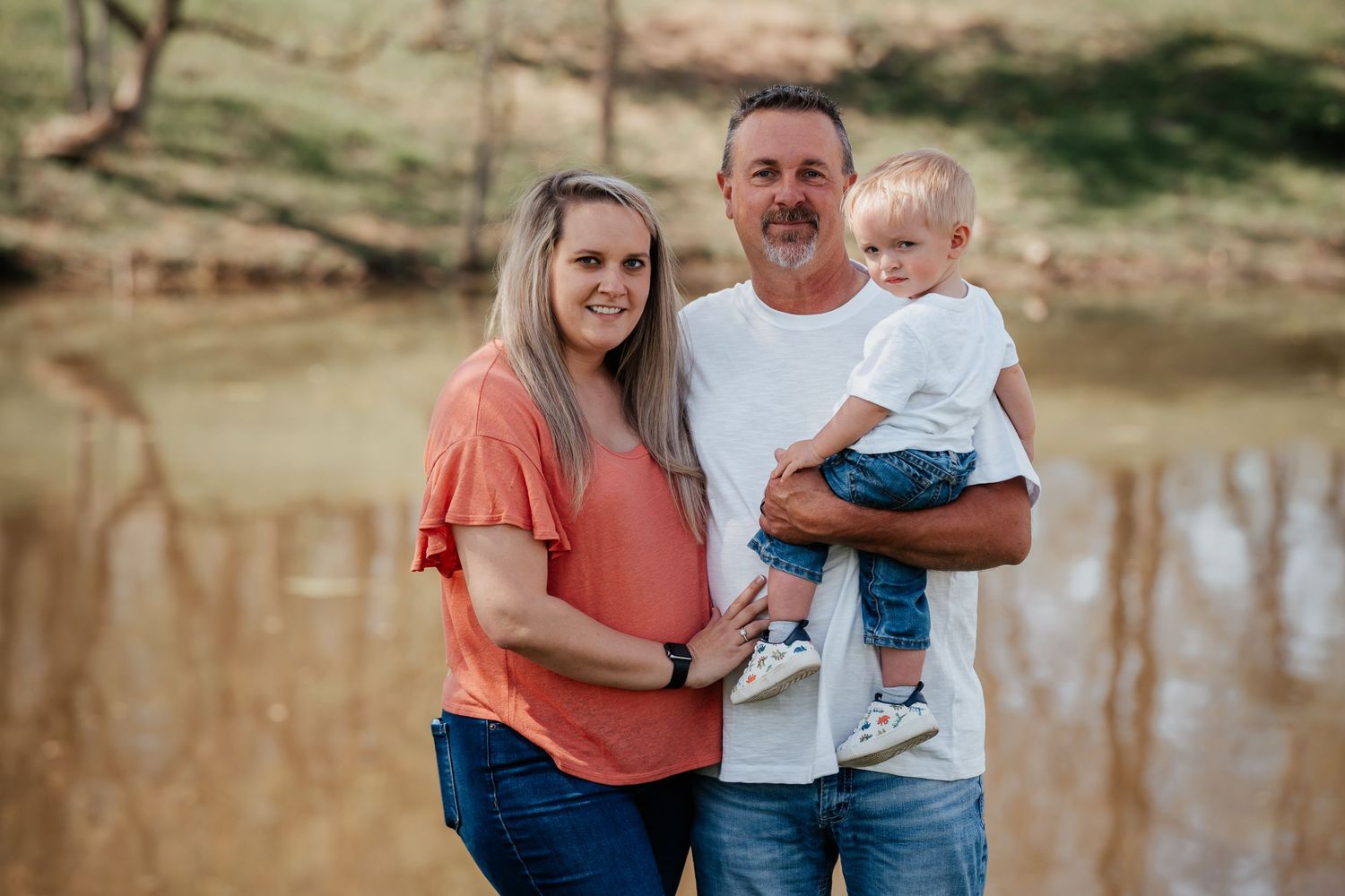 Family of three posed by pond during casual outdoor photo session.