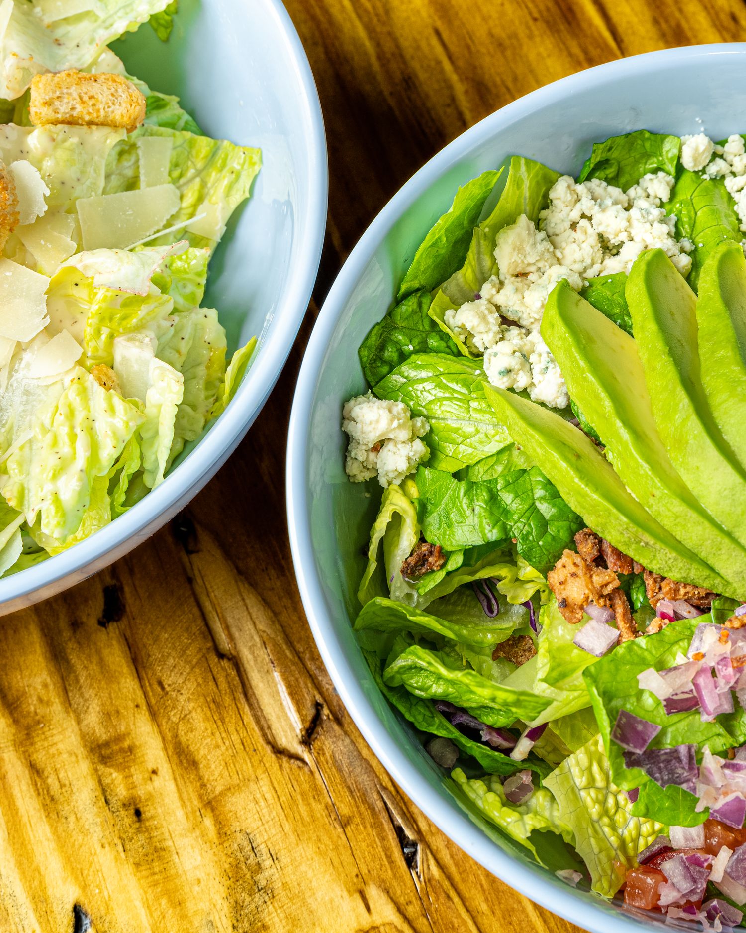 Two fresh salad bowls with lettuce, crumbled cheese and dressing on wooden table.