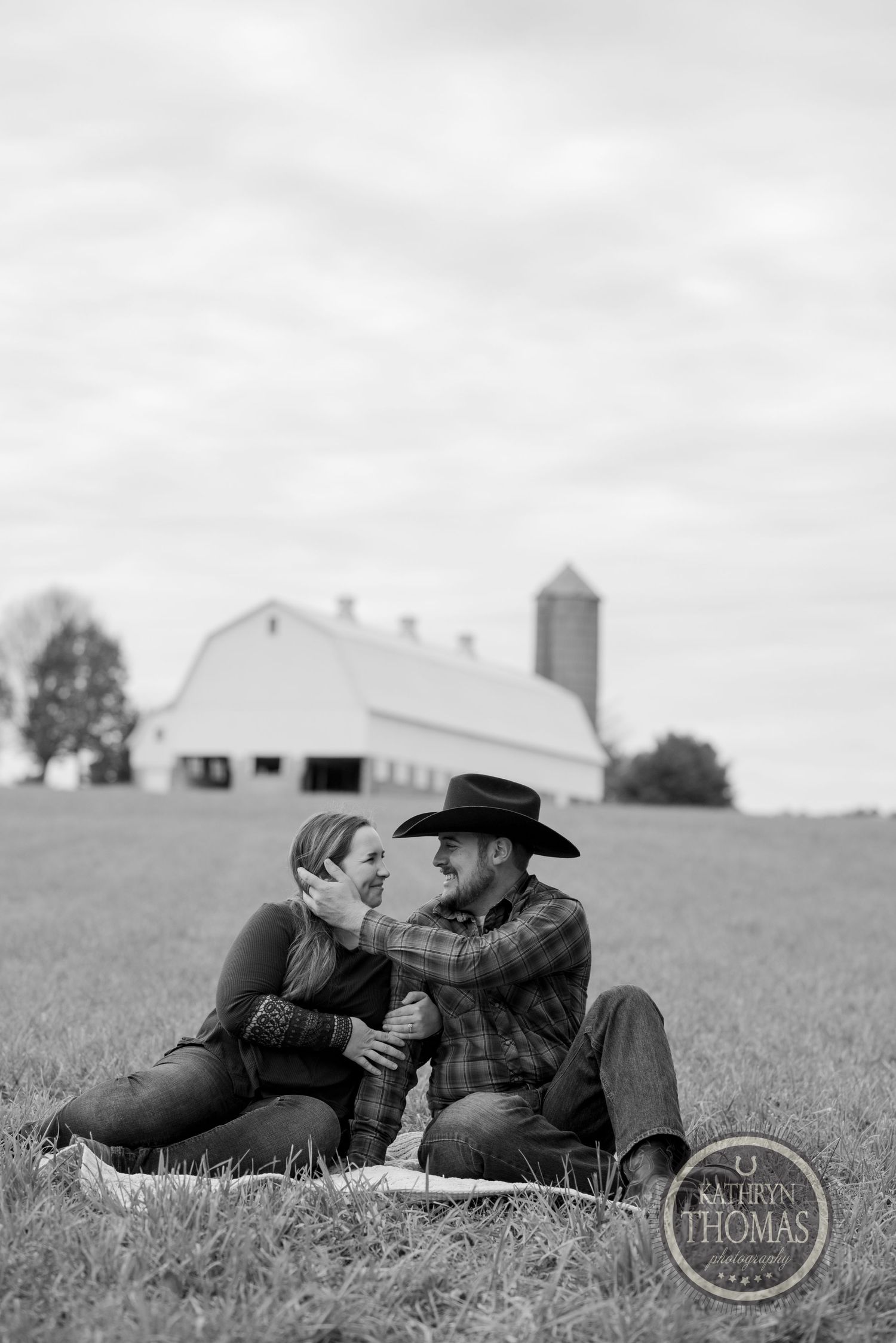 A romantic black and white portrait of a couple sitting together on a hill with a barn in the distance.