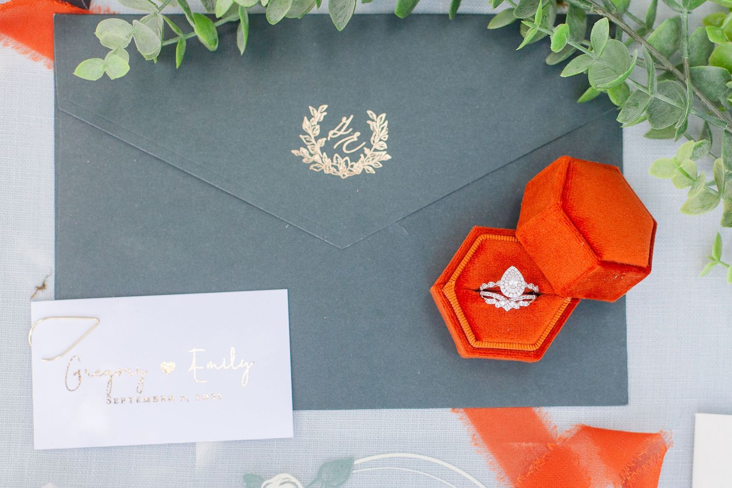 Wedding stationery with orange velvet ring box and gold monogram on gray envelope.
