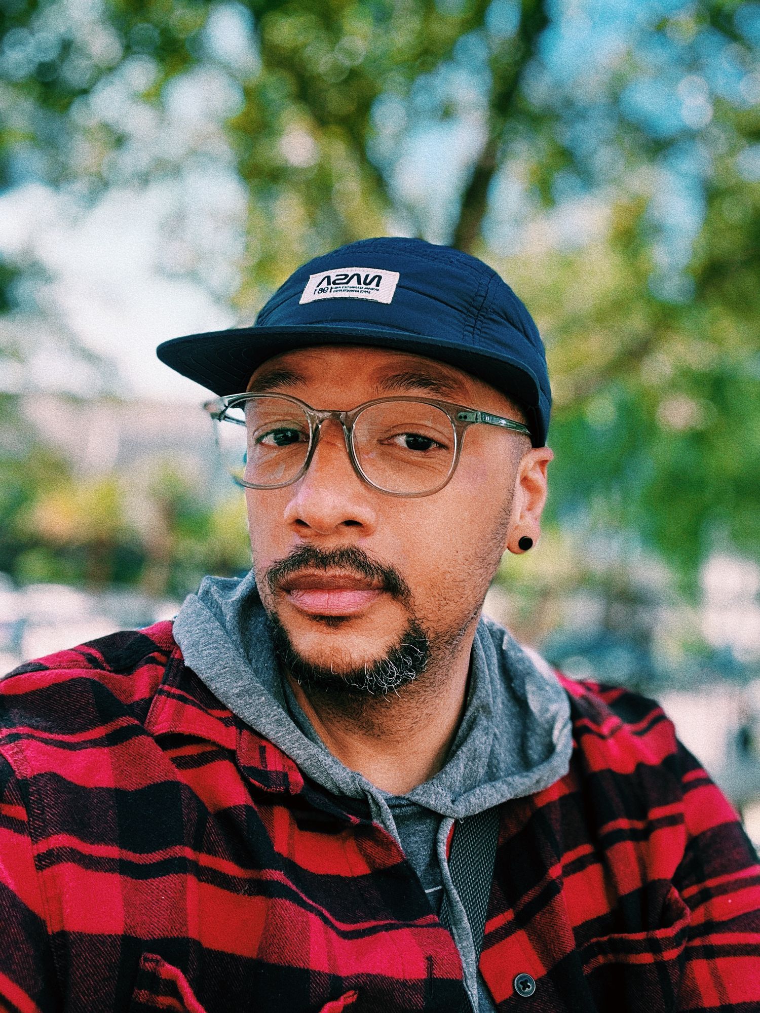 Portrait of someone wearing glasses, a navy cap and red plaid shirt against blurred green background.