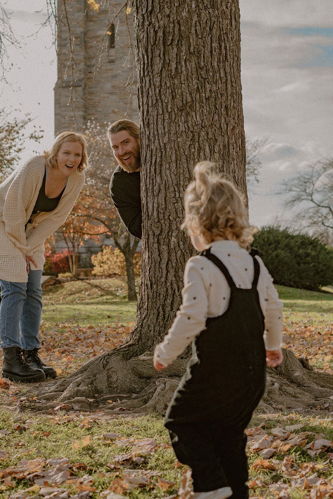 Parents playing peek-a-boo with toddler around large tree trunk in autumn setting.