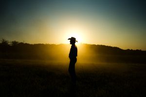 Silhouette of person in cowboy hat standing in field during dramatic sunset.