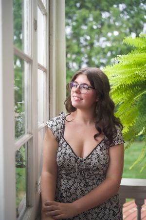 Portrait of woman in floral dress by window with fern plant in background.