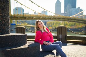Woman in bright pink sweater sitting by urban waterfront with city skyline in background.
