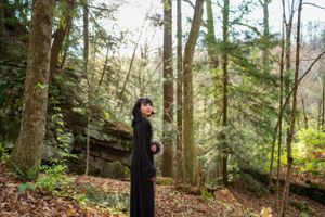 Figure in long black dress standing among tall trees in moody forest setting.