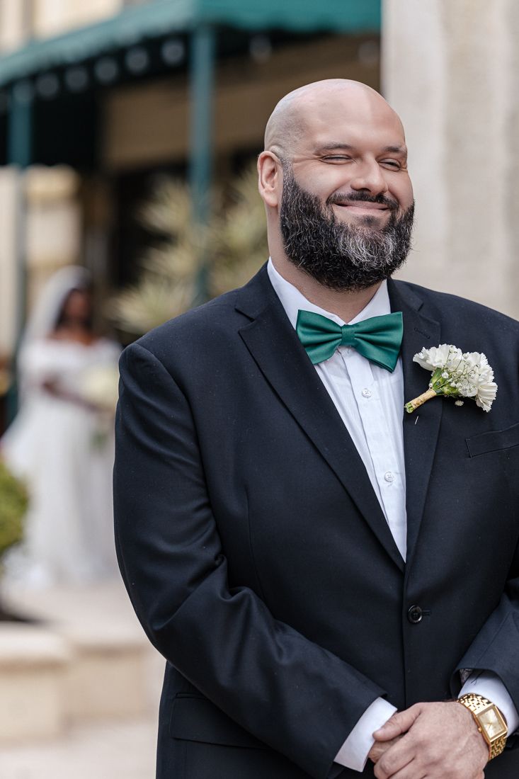 Wedding groom in classic tuxedo and emerald bow tie smiles while wearing floral boutonniere.