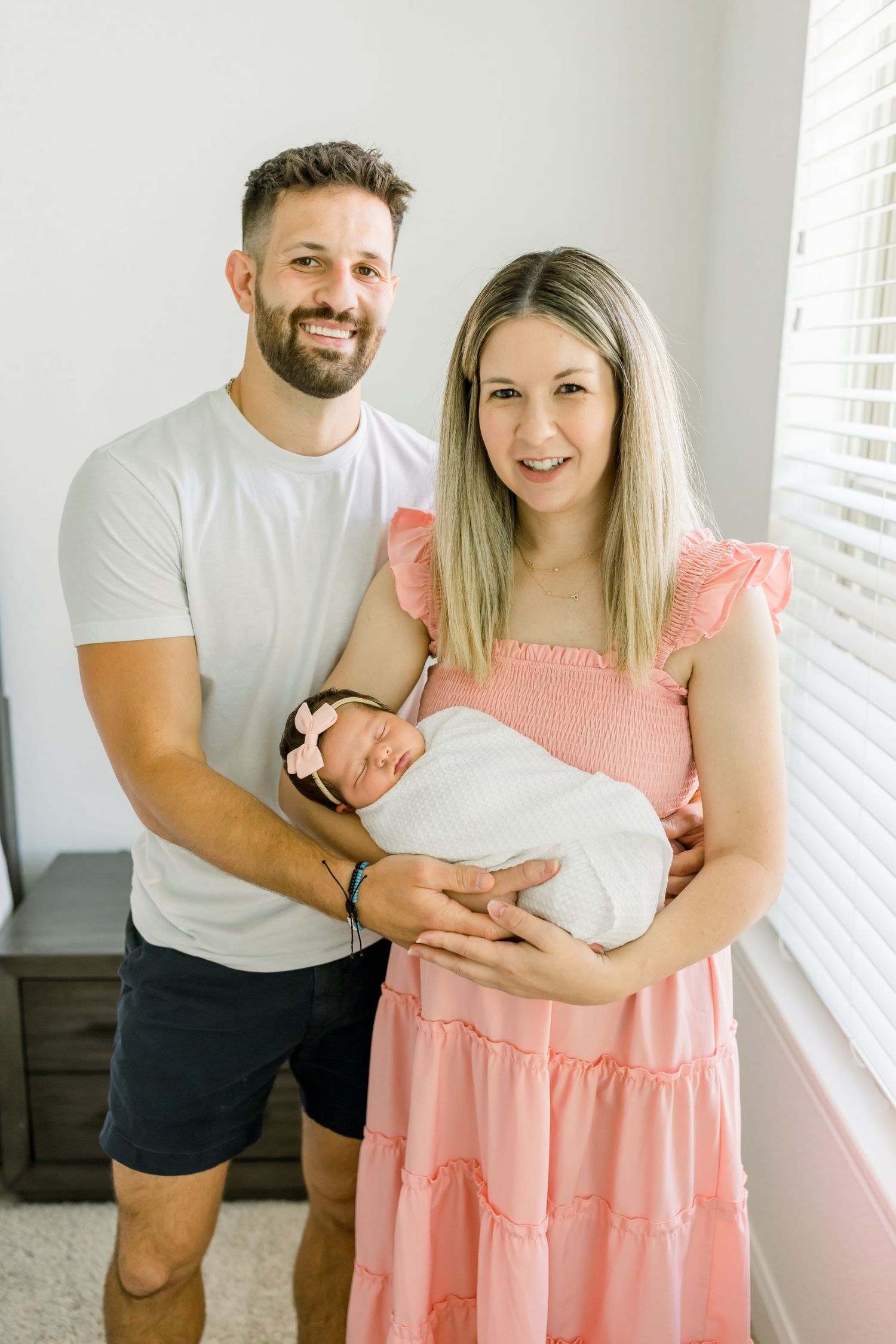 A couple holding their newborn baby while standing by a window in a minimalist room.