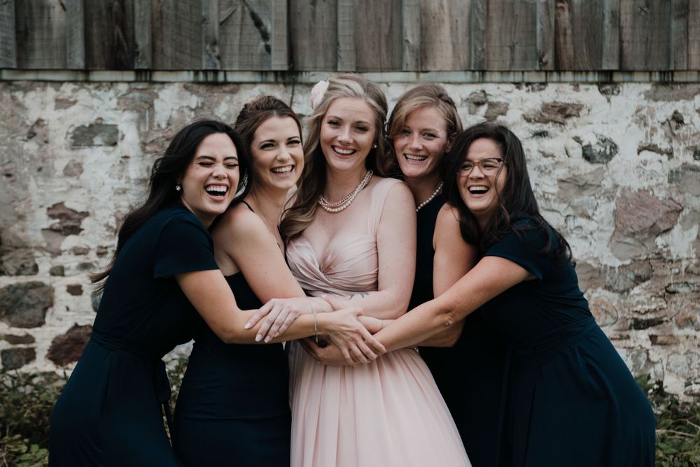 A bridal party in navy and blush dresses sharing a group hug against a stone wall.