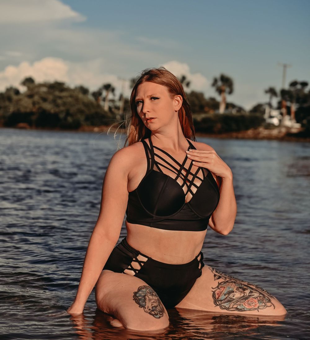 Figure in black strappy swimwear sitting in water with visible tattoos.