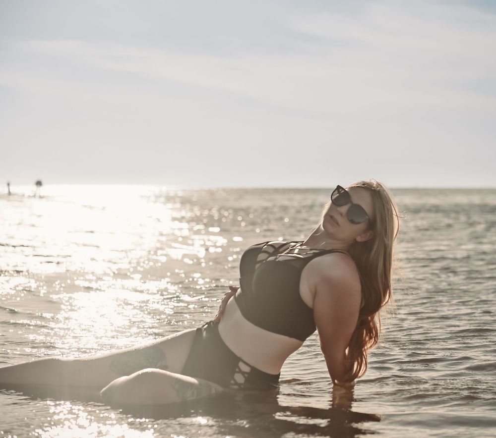 Athletic figure in black swimwear reclining in shallow ocean water at sunset.