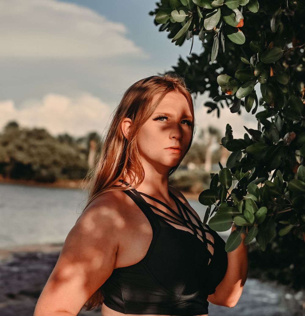 Athletic figure in black workout top poses against tropical foliage and waterfront backdrop during golden hour.