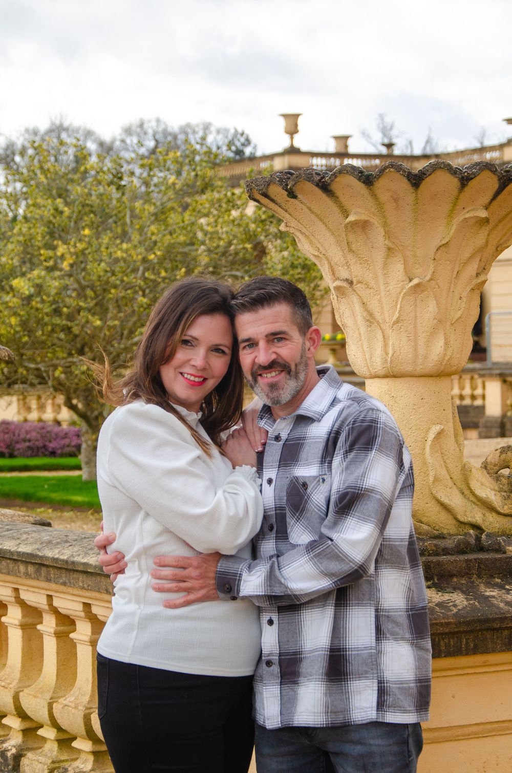 Couple embracing by ornate garden fountain with classical architecture in background.