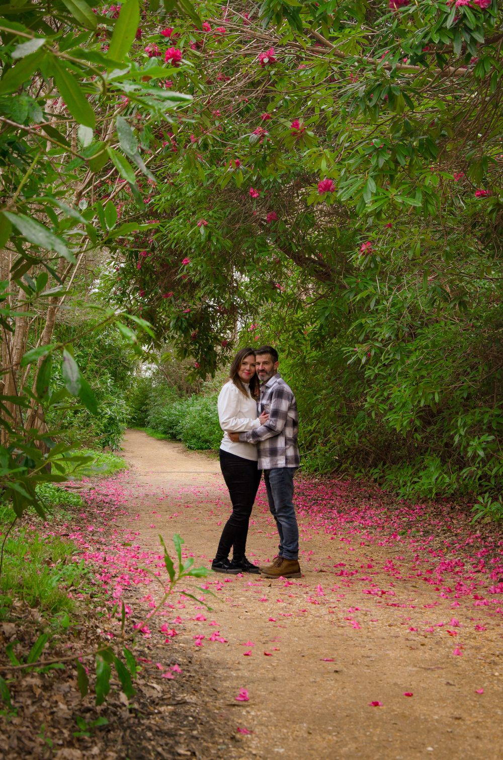 Couple standing on nature path with pink flowers scattered beneath trees.