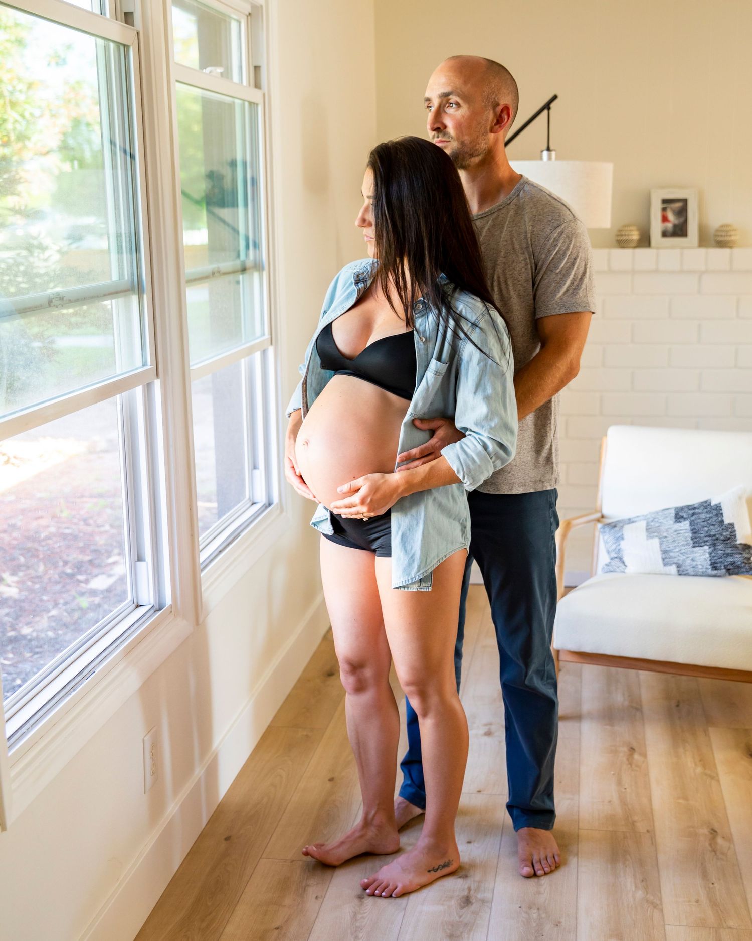 In-home maternity photoshoot with Expectant couple standing by window, man embracing pregnant woman from behind in cozy home setting.