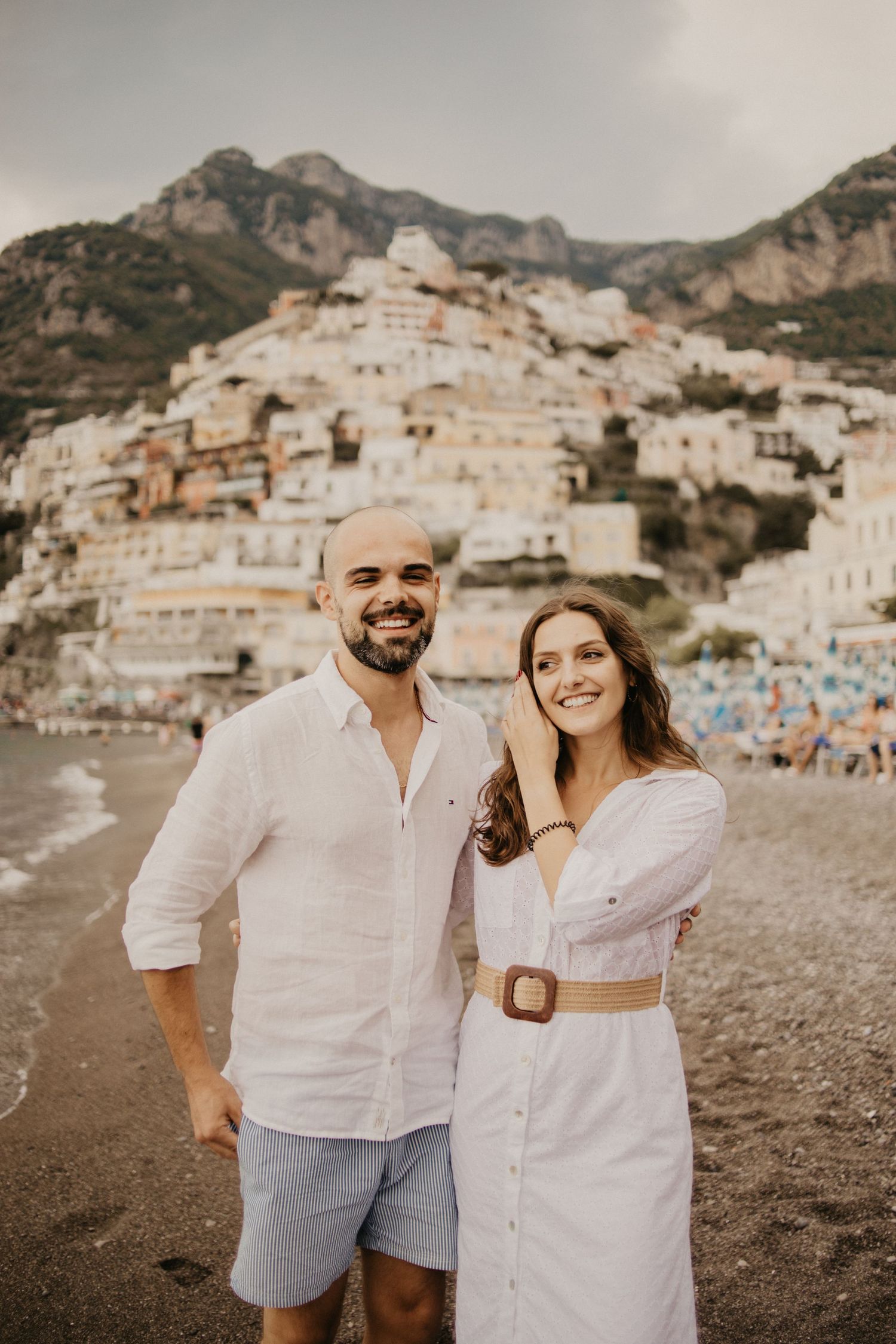 Happy couple in white outfits smiling on Positano beach with town vista behind them.