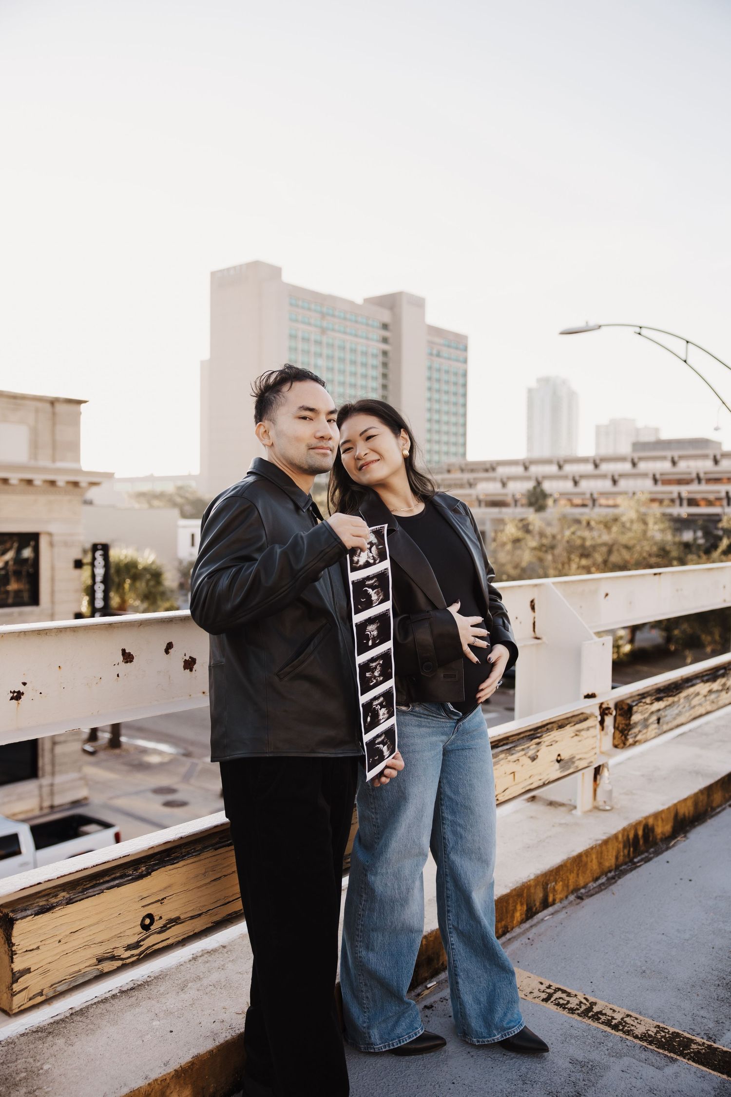 Expecting couple holding ultrasound photos on rooftop with city skyline in golden hour light.
