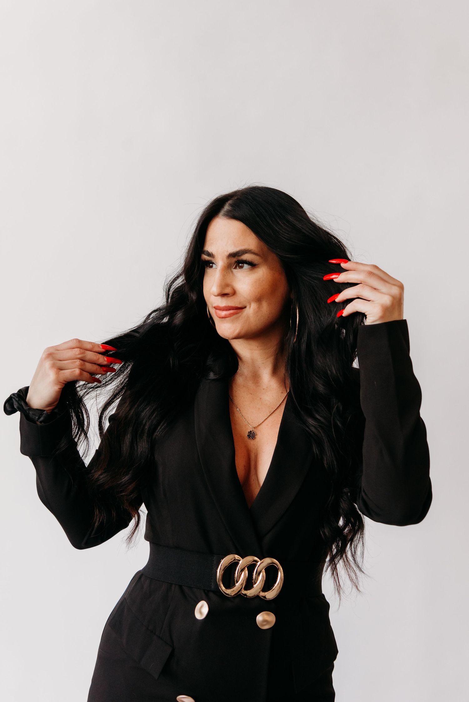 Woman in stylish black blazer with gold belt accessory poses against white background with long dark hair and red nails.
