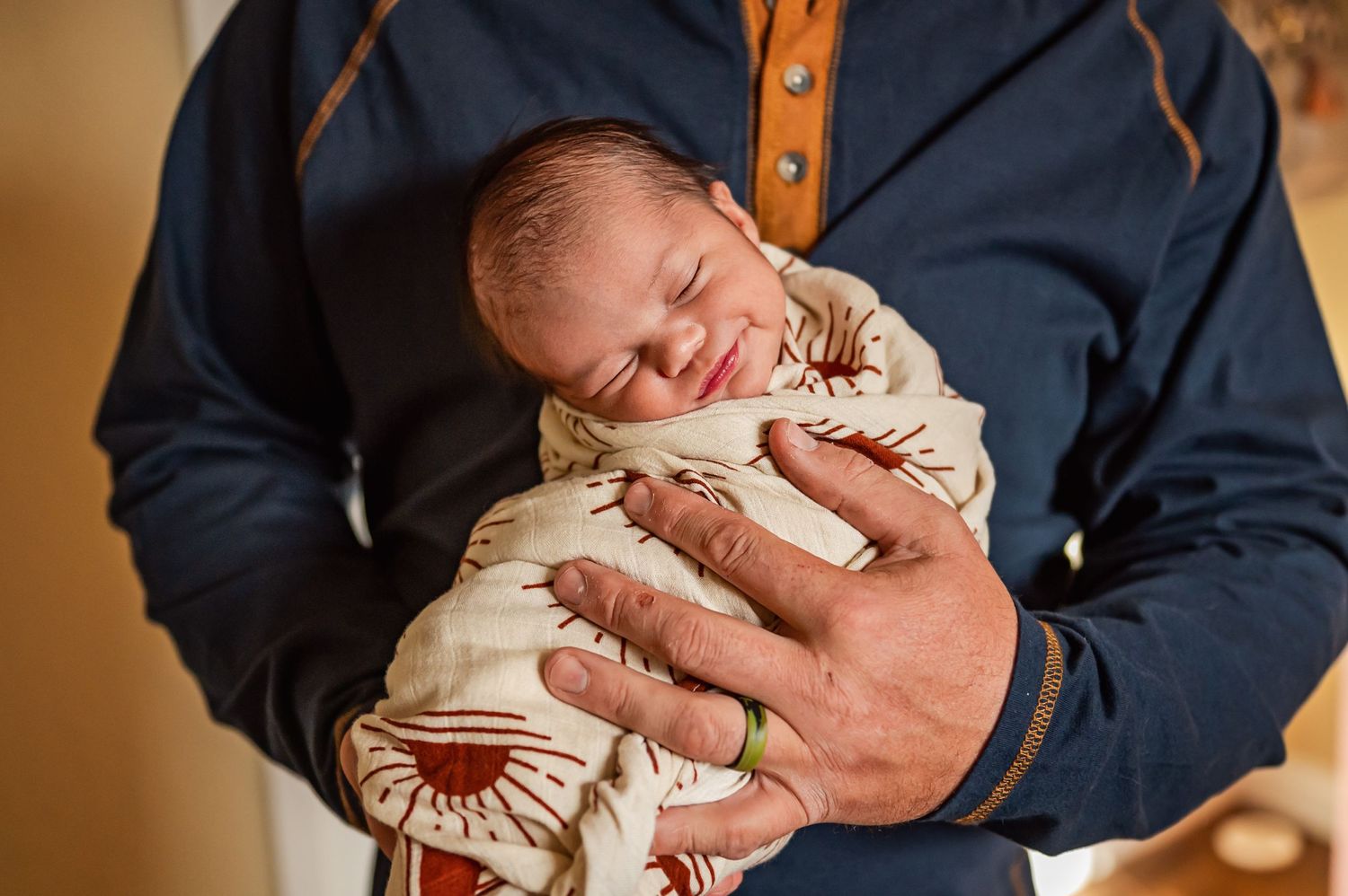 A sleeping newborn in cream-colored wrap is held gently against a navy blue shirt.