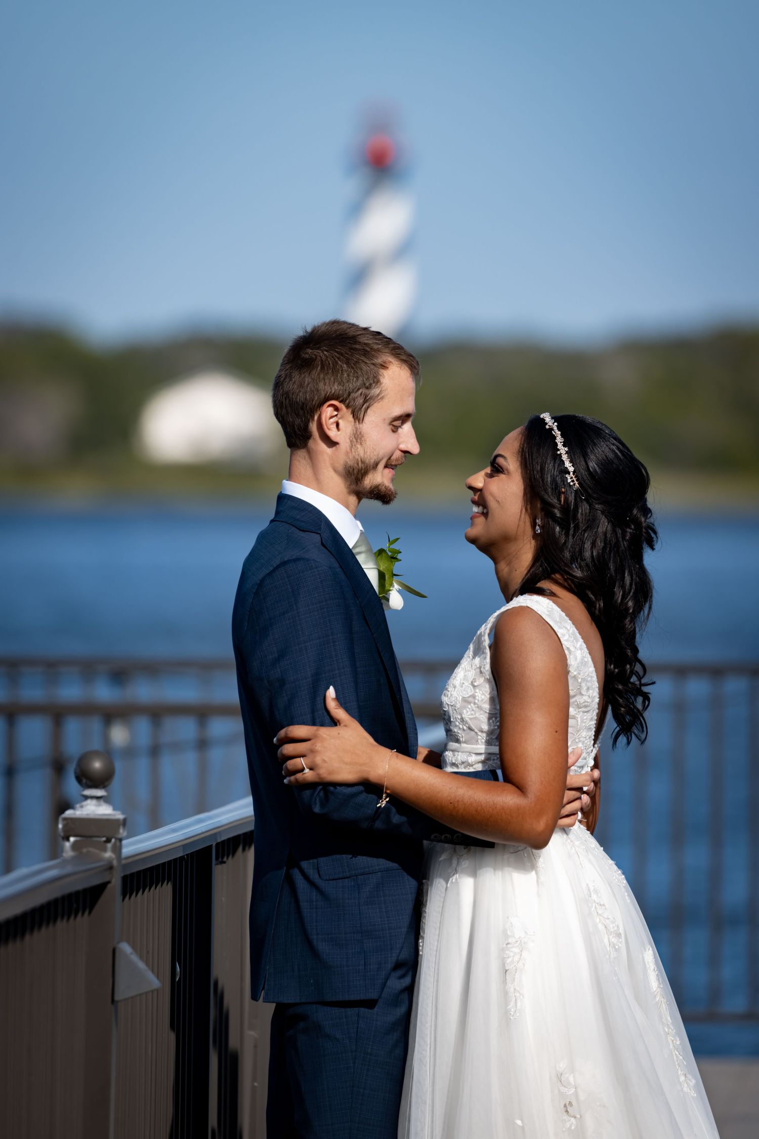 A couple shares a romantic moment on a pier with a lighthouse and waterfront view in the background.