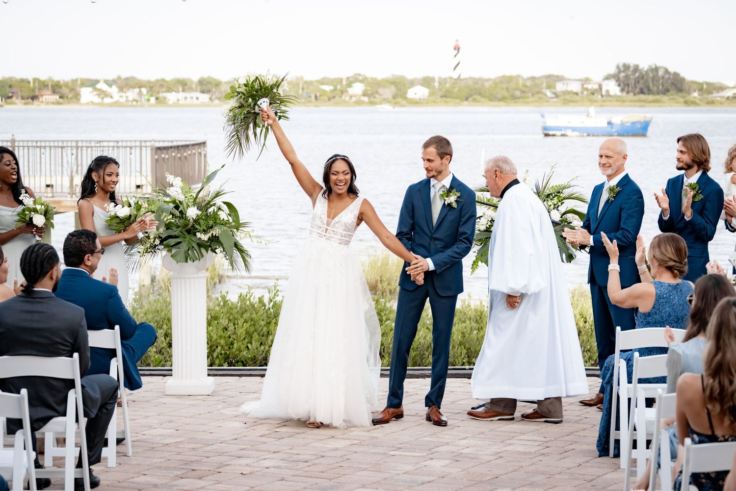 A waterfront wedding ceremony takes place with white chairs and floral arrangements by the water.