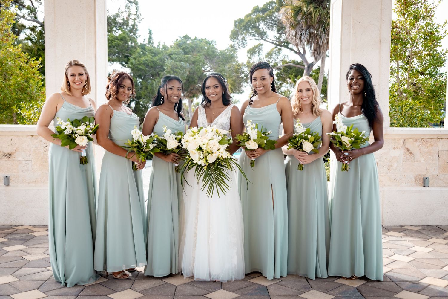 Bridesmaids in elegant sage green dresses hold white and green bouquets while posing at an outdoor venue.