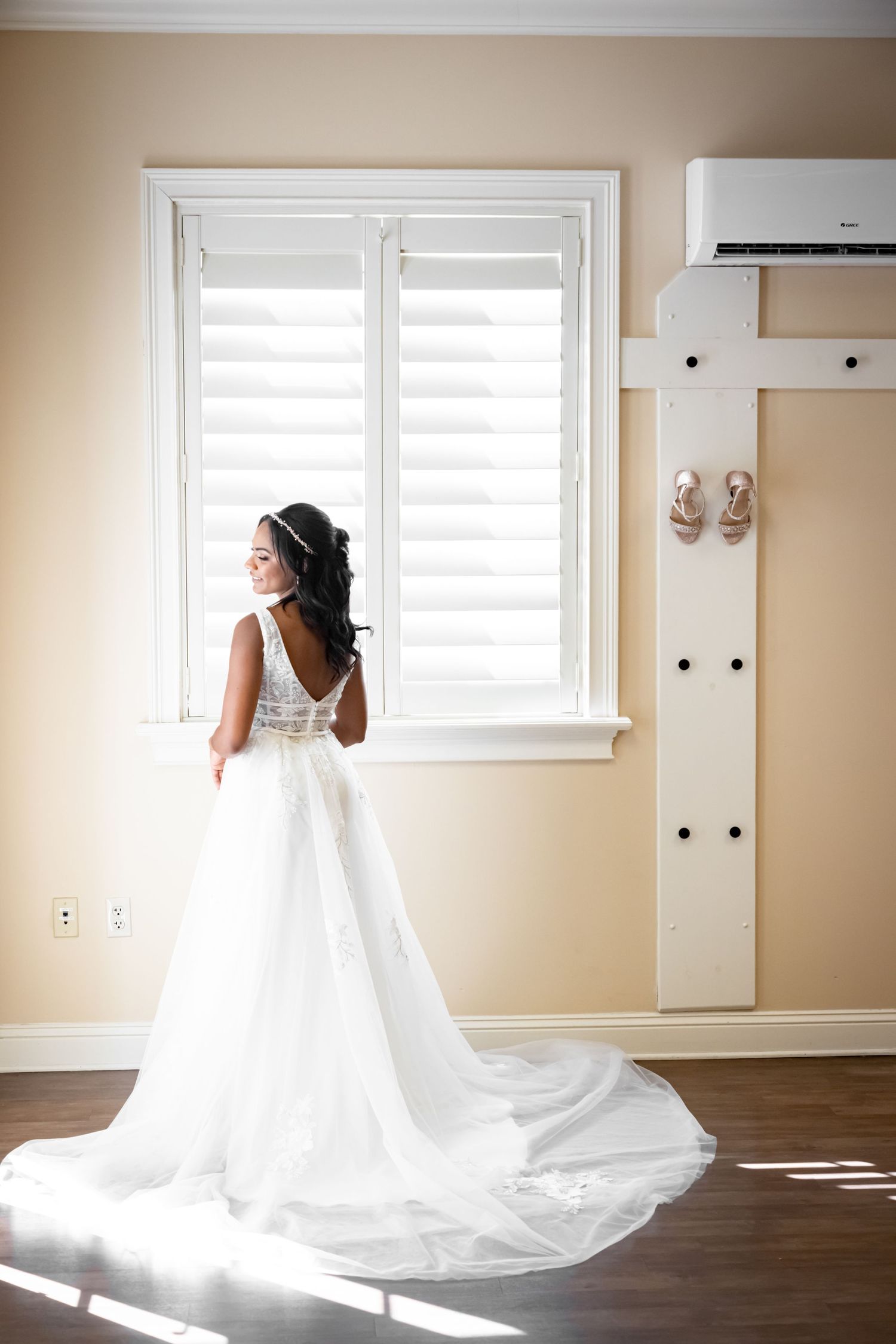 A white wedding gown with a flowing train and open back stands illuminated by natural window light.