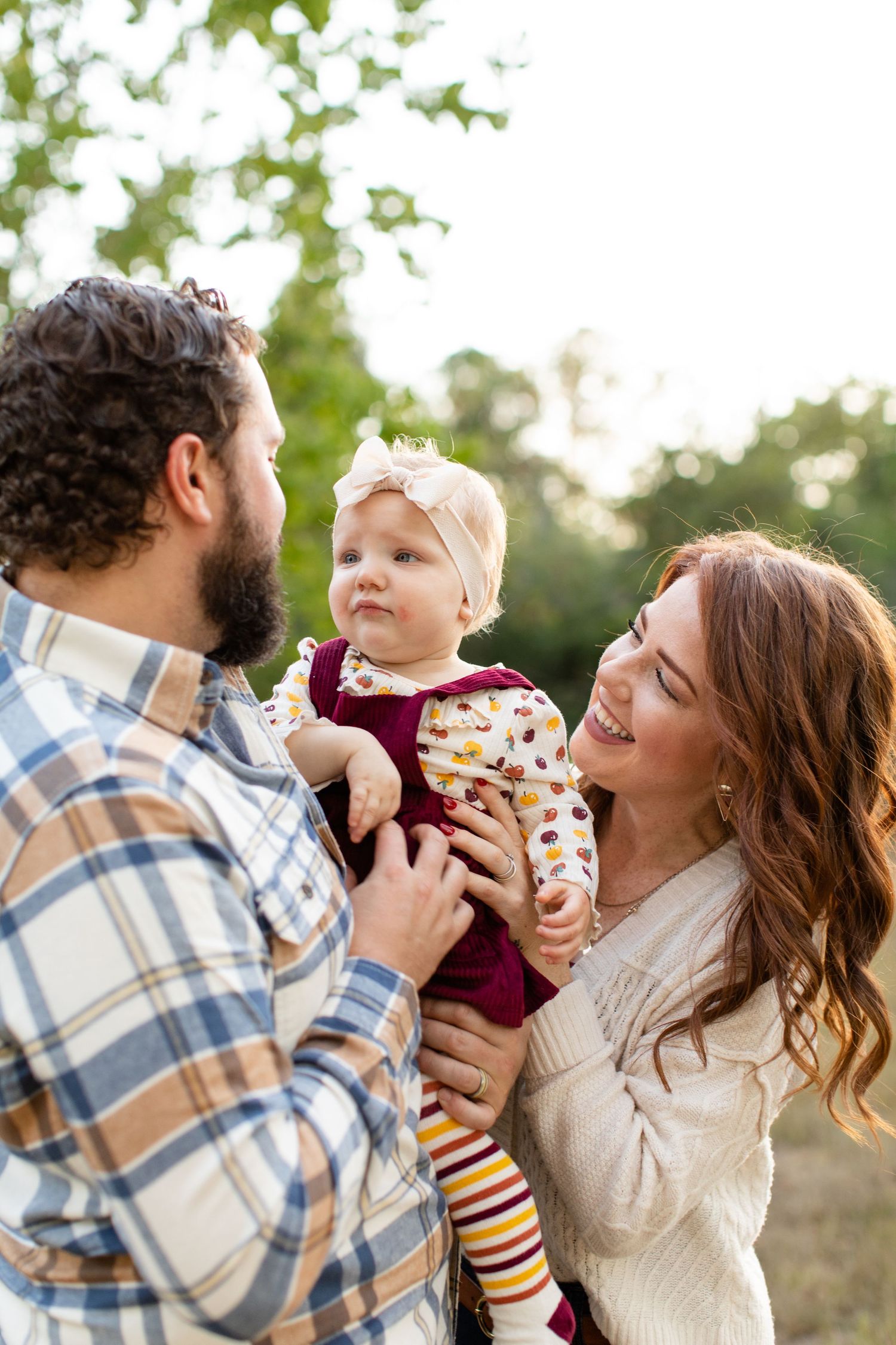 Candid outdoor family moment with toddler in burgundy dress being held between parents in fall clothing.